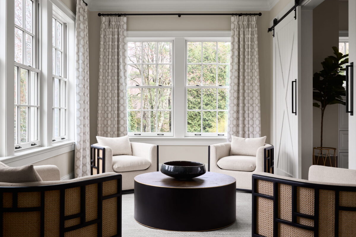 Bright living room with large windows, two cream chairs, a round dark coffee table, and patterned curtains. The space has light walls, sliding barn doors, and modern, minimalist decor.