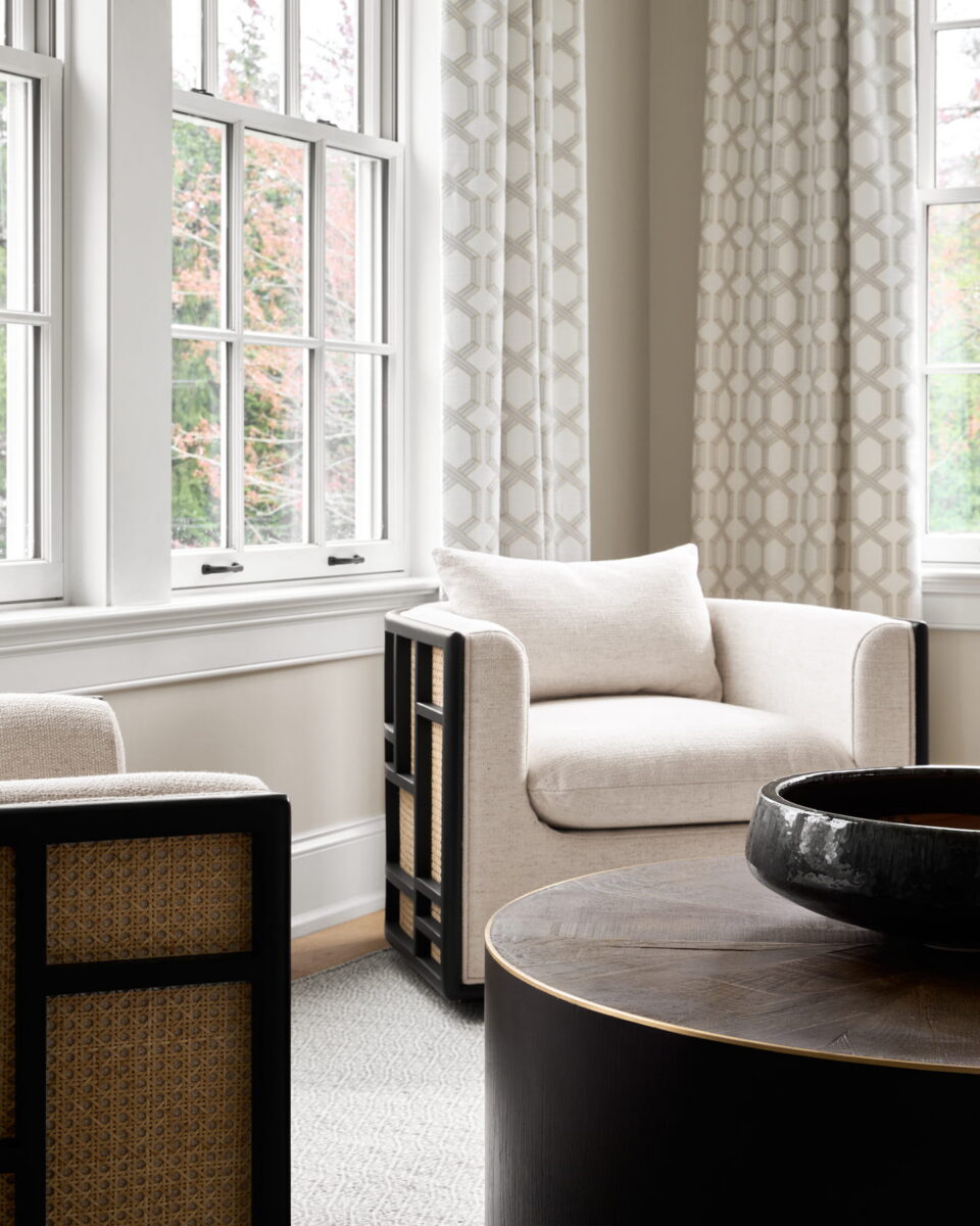 A bright living room with large windows, cream-colored armchairs, geometric-patterned curtains, and a round dark wood coffee table with a black bowl. Light filters through, creating a cozy, modern atmosphere.
