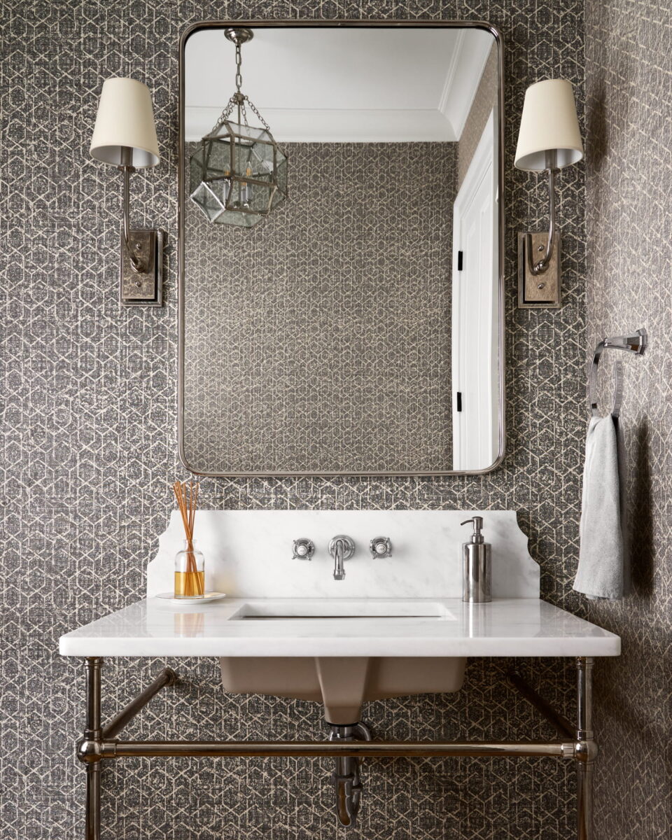 Elegant bathroom with patterned wallpaper, marble sink, metal fixtures, a large rectangular mirror, two wall sconces, a hanging light, a hand towel, and a soap dispenser on the counter.