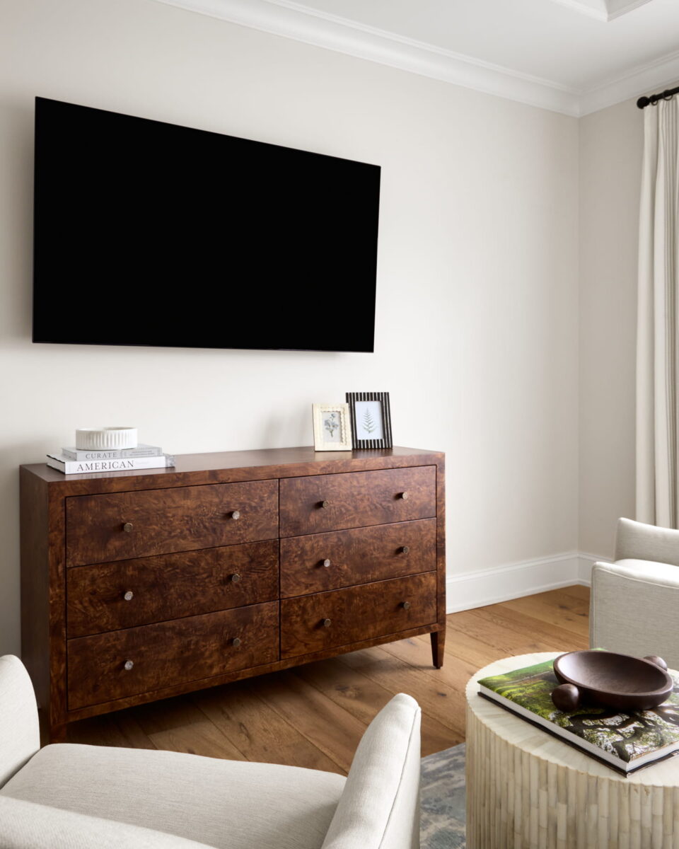 A modern living room with a large black TV mounted above a wooden dresser, books and framed photos on top, two light-colored chairs, wood flooring, and white curtains.