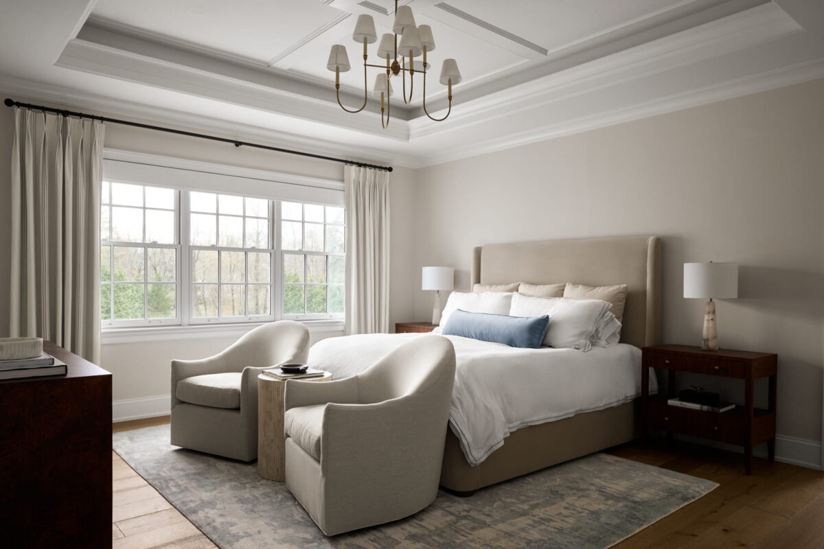 A modern bedroom with a large bed, two beige armchairs at the foot, a blue accent pillow, nightstands, lamps, a chandelier, and a large window with curtains letting in natural light.