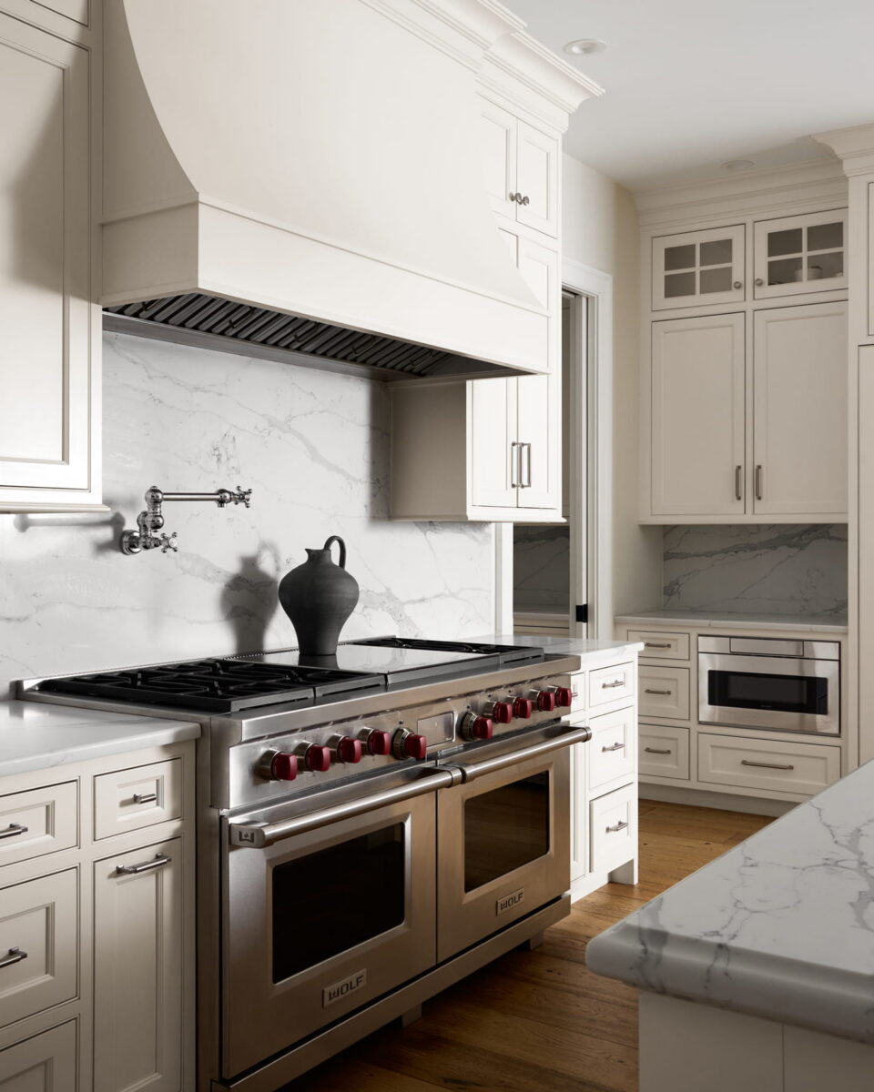 Modern kitchen with white cabinets, marble backsplash, stainless steel double oven, red burner knobs, and a black vase on the stovetop. Light wood floor and built-in microwave visible.