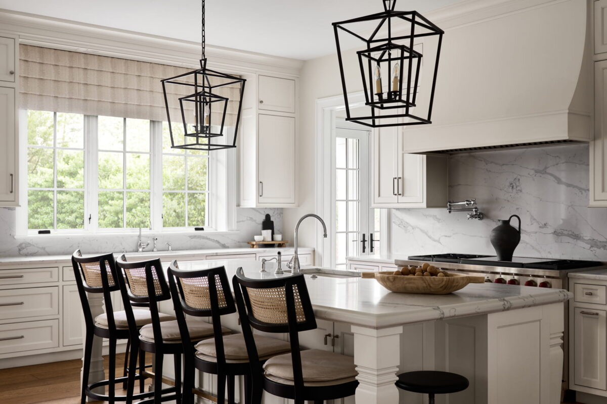 Modern white kitchen with marble countertops, a central island with four black chairs, large pendant lights, a window with a beige shade, and a bowl of fruit on the counter.