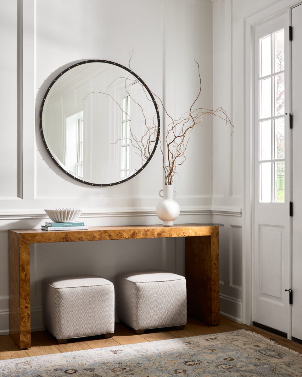 A modern entryway with a wooden console table, two white ottomans, a round mirror, a white vase with curly branches, a decorative bowl, and a door with glass panels.