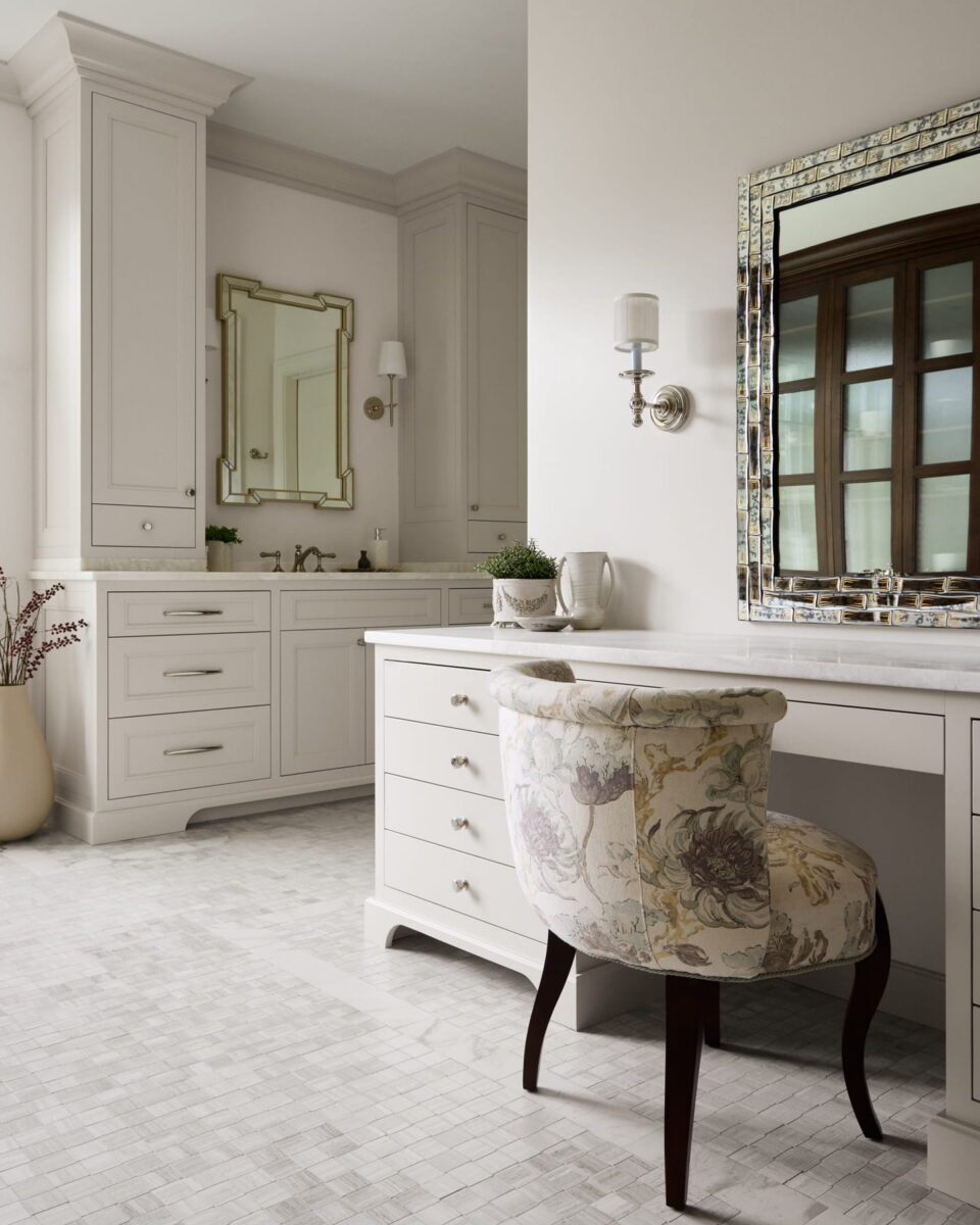 Elegant bathroom with a white vanity, floral upholstered chair, large mirror with mosaic frame, and a second sink area. Neutral tones, tile floor, and decorative vase add a luxurious, serene atmosphere.