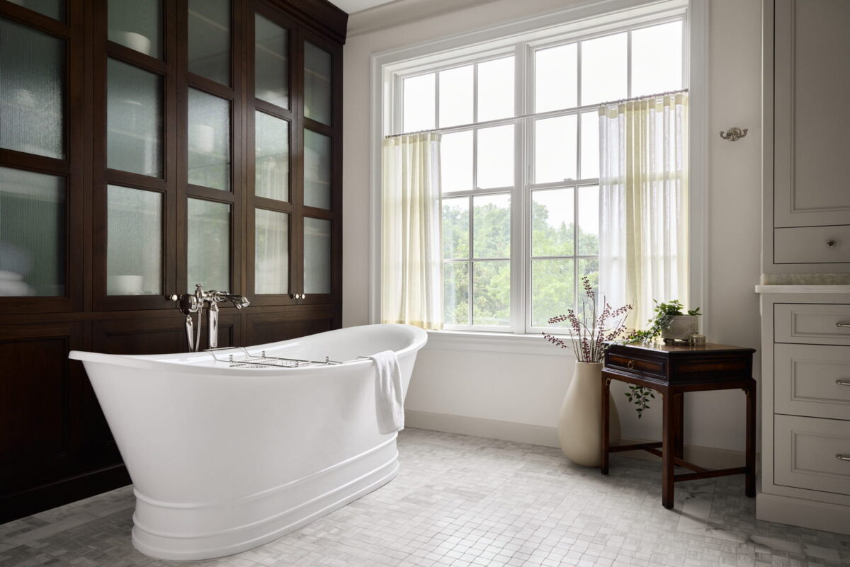 Bright bathroom with a white freestanding tub, wooden built-in cabinets, a large window with sheer curtains, and a small table with a vase of greenery.