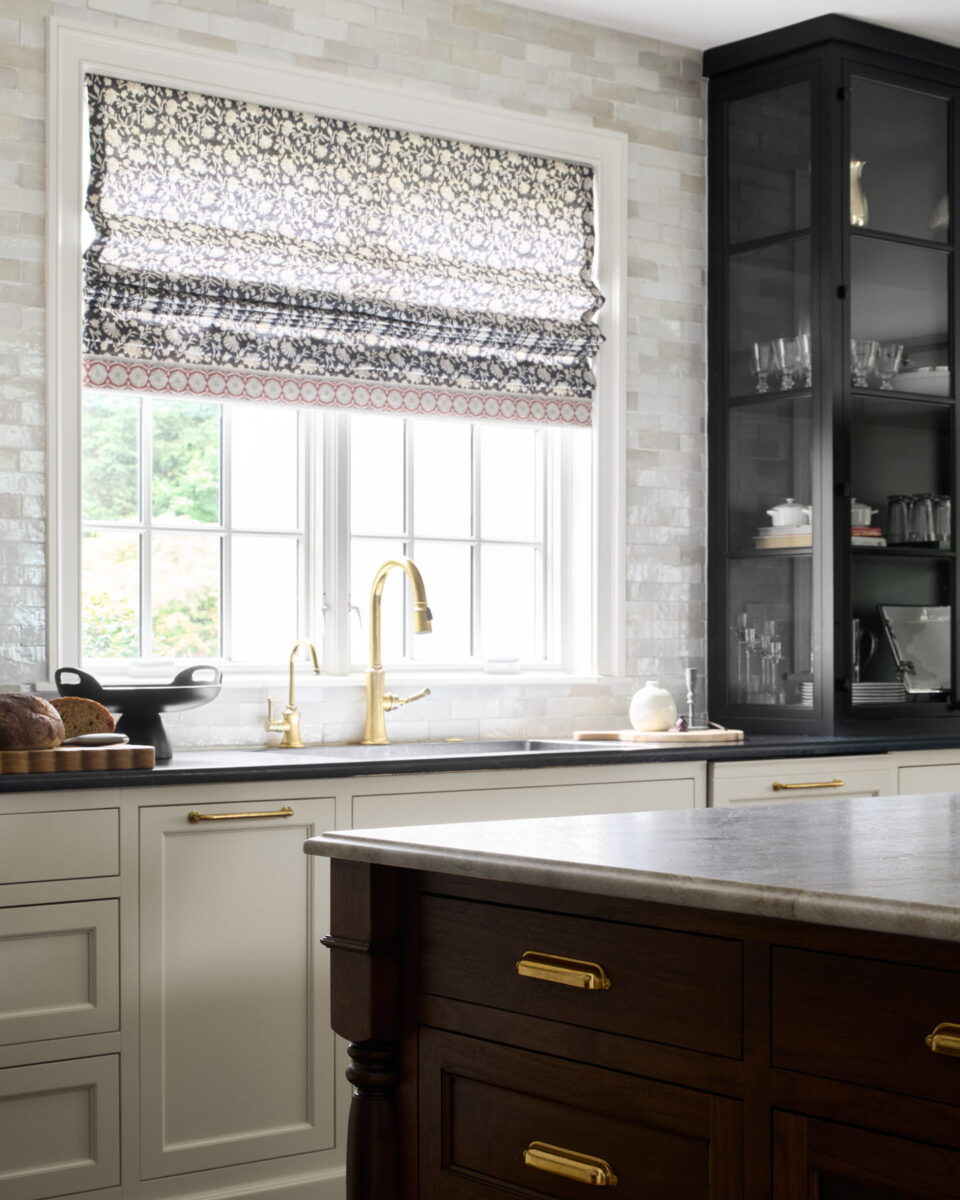 A bright kitchen with white cabinets, gold hardware, a marble countertop, and a patterned roman shade on the window above a gold faucet. Glassware is displayed in a black cabinet.