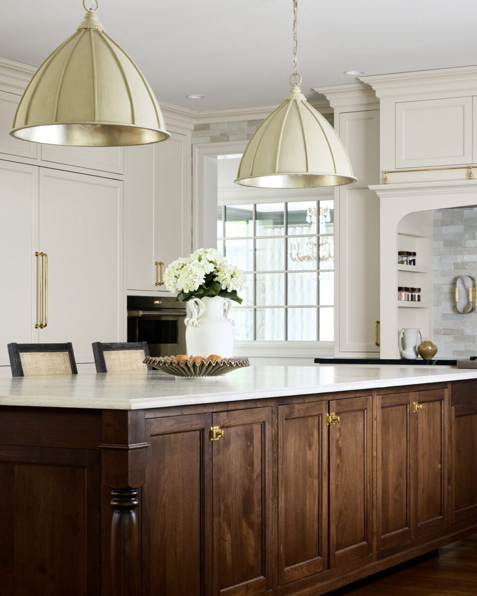 Elegant kitchen with white cabinets, a large dark wood island, marble countertop, gold pendant lights, and a white vase with flowers. Warm lighting and a bright, airy atmosphere complete the sophisticated design.