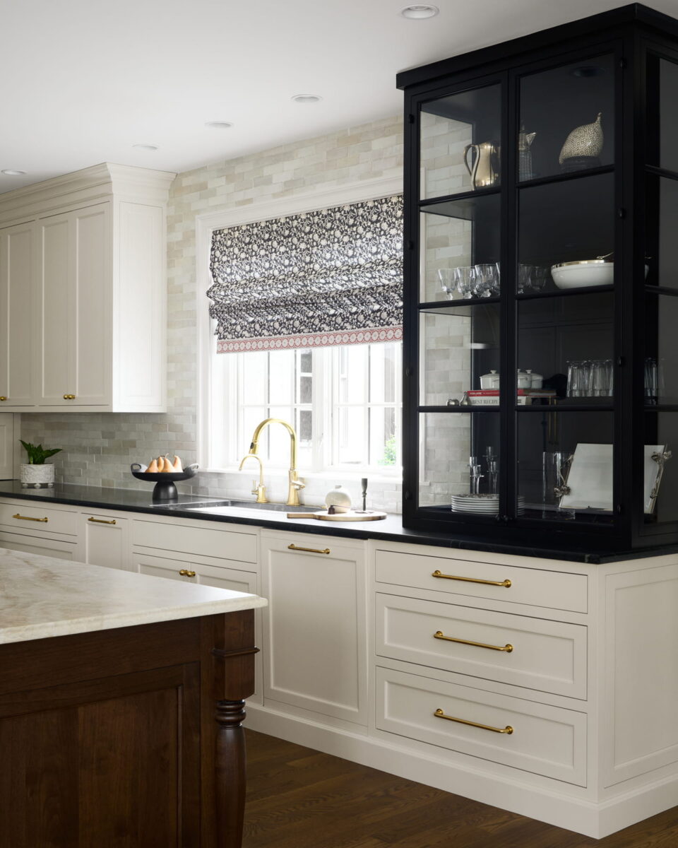 Bright kitchen with white cabinets, gold hardware, marble countertop, black glass display cabinet, patterned Roman shade over window, and wood island on a dark wood floor. Various kitchen items are displayed on the counter and shelves.
