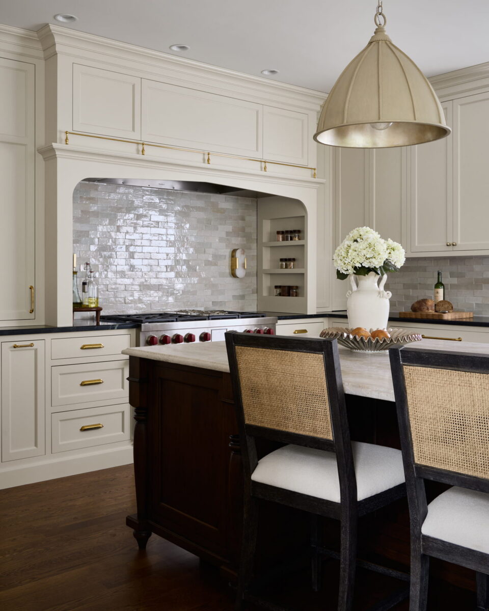 Elegant kitchen with cream cabinets, gold hardware, a marble island, dark wood accents, a large pendant light, and a vase of white flowers. Glossy tile backsplash and built-in shelves add a sophisticated touch.