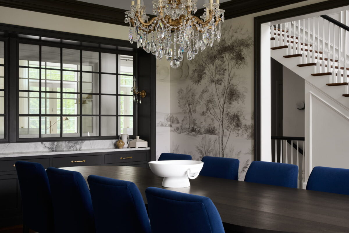 Elegant dining room with a dark wood table, blue velvet chairs, a crystal chandelier, black cabinetry, and scenic wallpaper. A white bowl sits in the center of the table. A staircase is visible in the background.