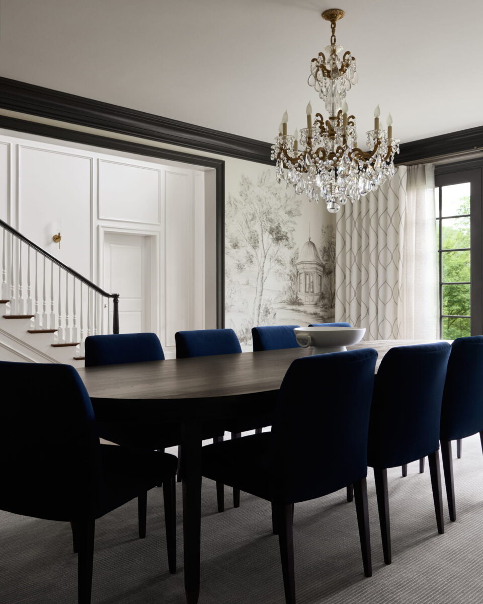 Elegant dining room with a dark wood table, eight blue chairs, a crystal chandelier, large windows with sheer curtains, and a staircase in the background. Wall art features a detailed black and white landscape.