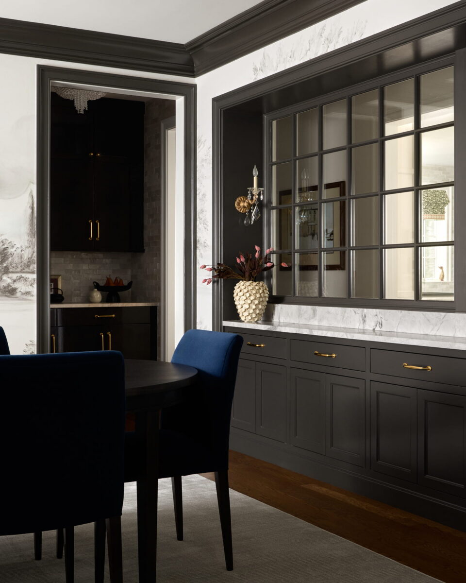 Dining area with a black table, blue chairs, and built-in black cabinetry with gold handles. A decorative pineapple vase and wall sconce sit on a marble countertop under a large window.
