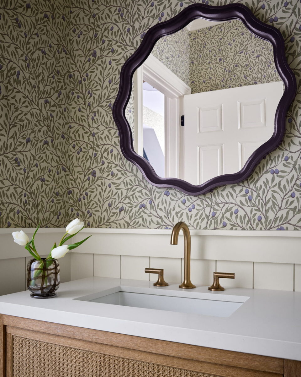 A bathroom with leafy wallpaper, a wavy-edged round mirror, brass faucet and handles, a white countertop, wooden vanity, and a glass vase with white tulips.