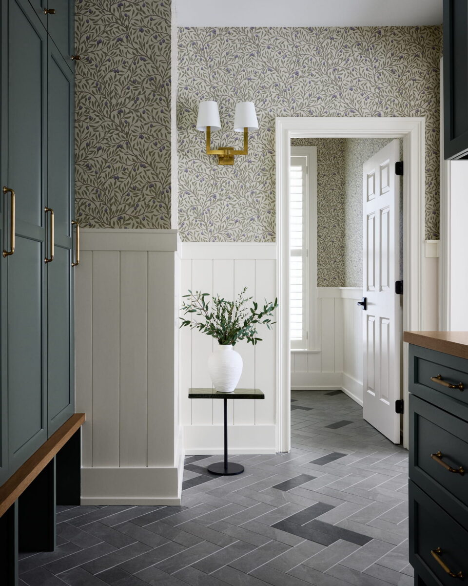 Mudroom with green cabinets, wood accents, white wainscoting, patterned wallpaper, gray tile floor, a white vase with greenery on a black table, and a wall sconce above. Doorway opens to a sunlit room with shutters.