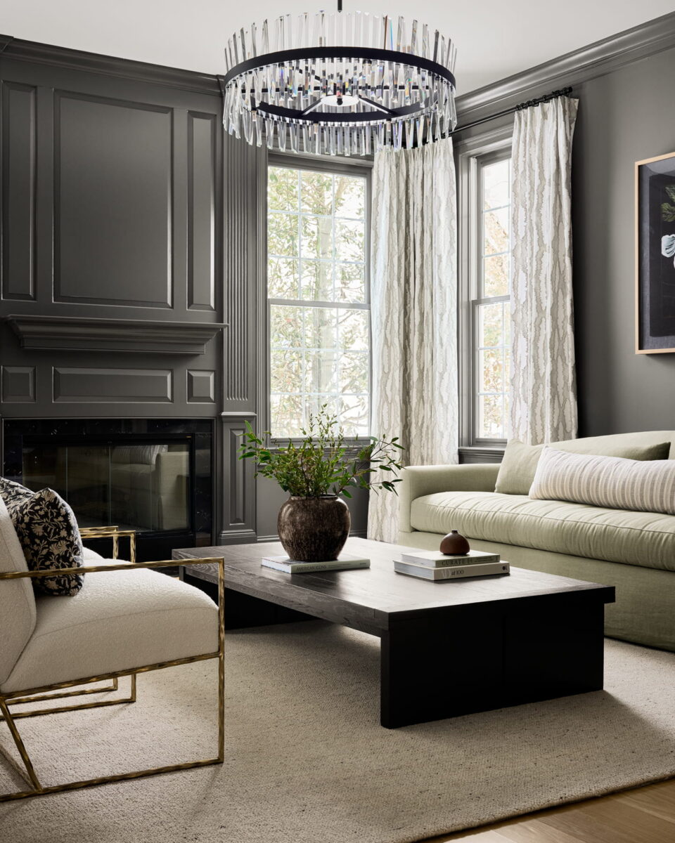A modern living room with dark paneled walls, a fireplace, large windows with patterned curtains, a light sofa, a white armchair, a dark coffee table with books and a vase, and a crystal chandelier above.