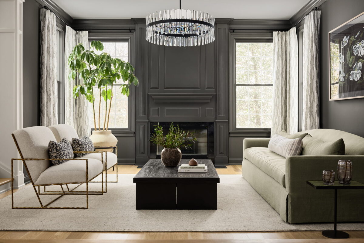 Modern living room with dark gray walls, large windows, a light area rug, a green sofa, a light armchair, a black coffee table with decor, a potted plant, and a crystal chandelier.