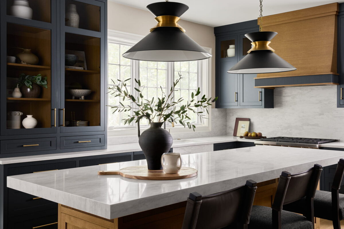 Modern kitchen with dark blue cabinets, marble countertops, gold-accented black pendant lights, and a wooden island. A vase with greenery and a cup sit on a round tray. Sunlight streams through a large window.