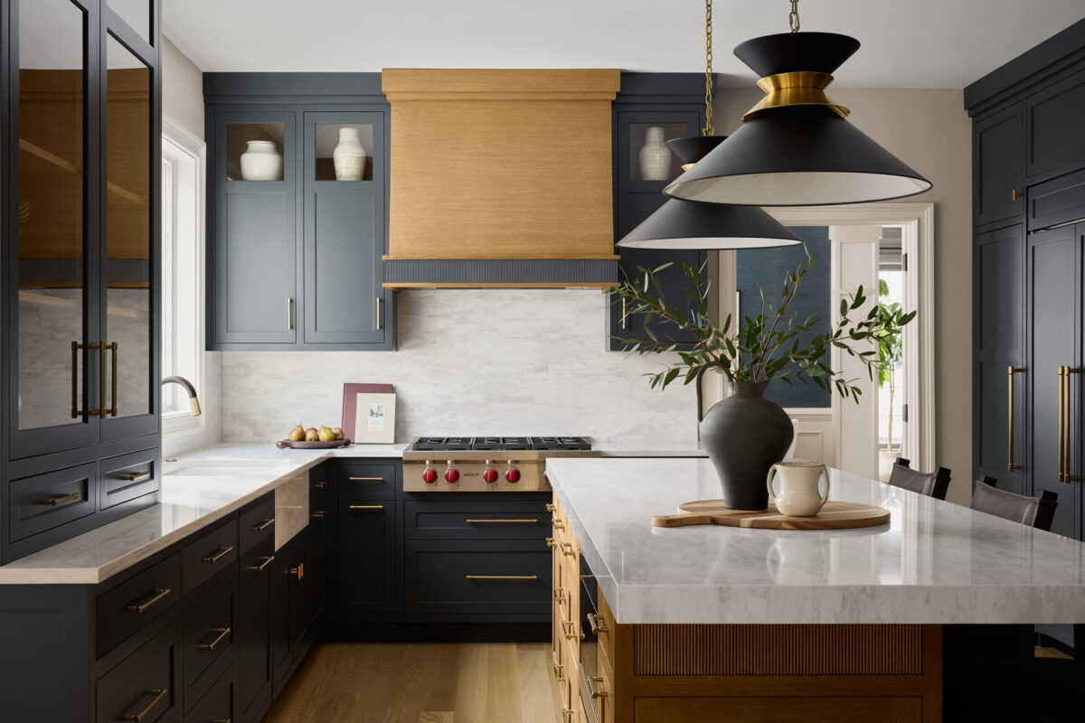 Modern kitchen with navy cabinets, brass hardware, white marble countertops, wooden accents, a stovetop, pendant lights, and a vase with greenery on a large island.