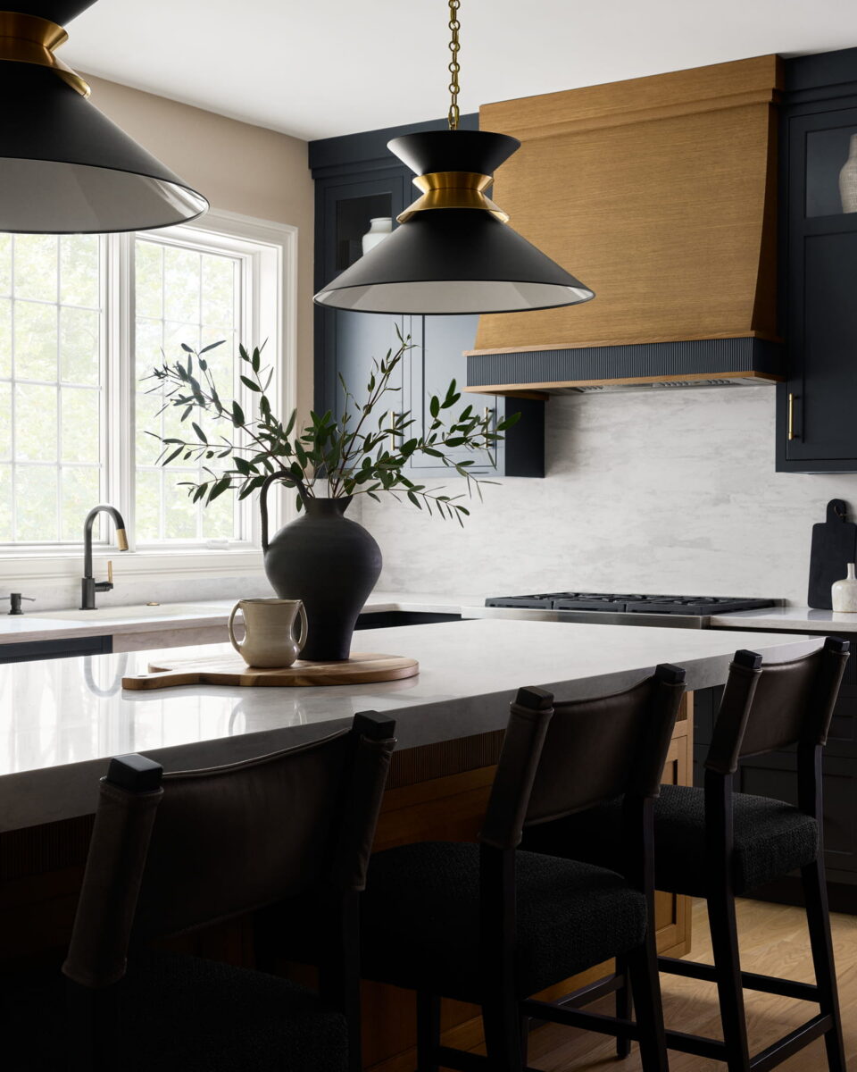 Modern kitchen with black chairs at a white island, black and gold pendant lights, a vase with greenery, and a window letting in natural light above the sink.
