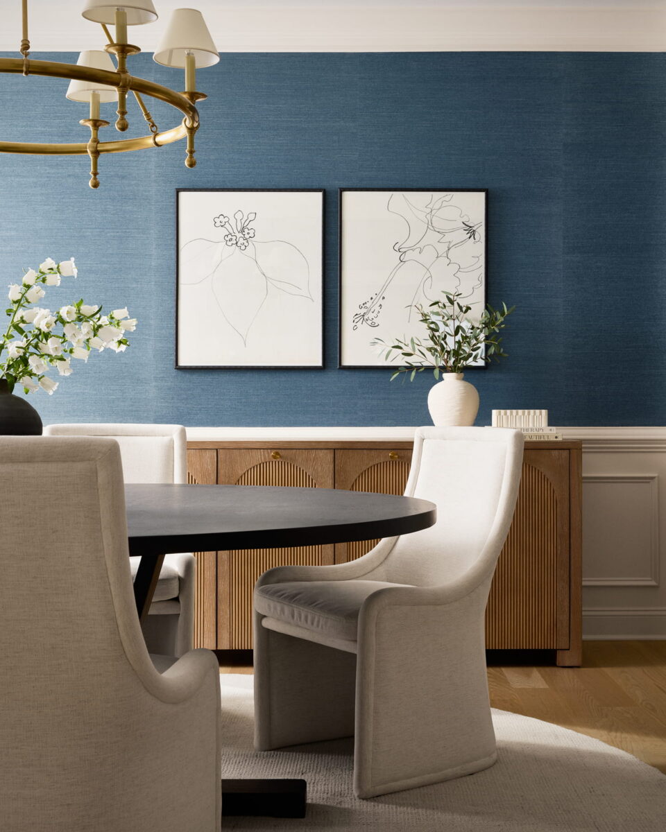A modern dining room with a round black table, light upholstered chairs, wooden sideboard, blue textured wall, two minimalist art pieces, and a gold chandelier. A white vase with flowers sits on the sideboard.