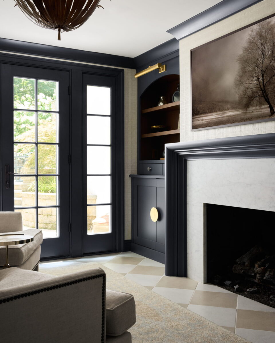 Modern living room with black French doors, marble fireplace, beige armchair, built-in shelves, and framed landscape art above the mantel; sunlight streams in from outside.