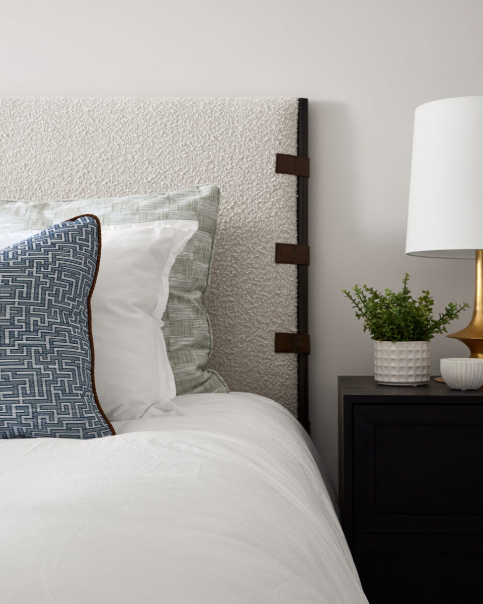 A bed with white and green pillows, a white textured headboard, and a black nightstand holding a gold lamp, a small plant in a white pot, and a round striped container.