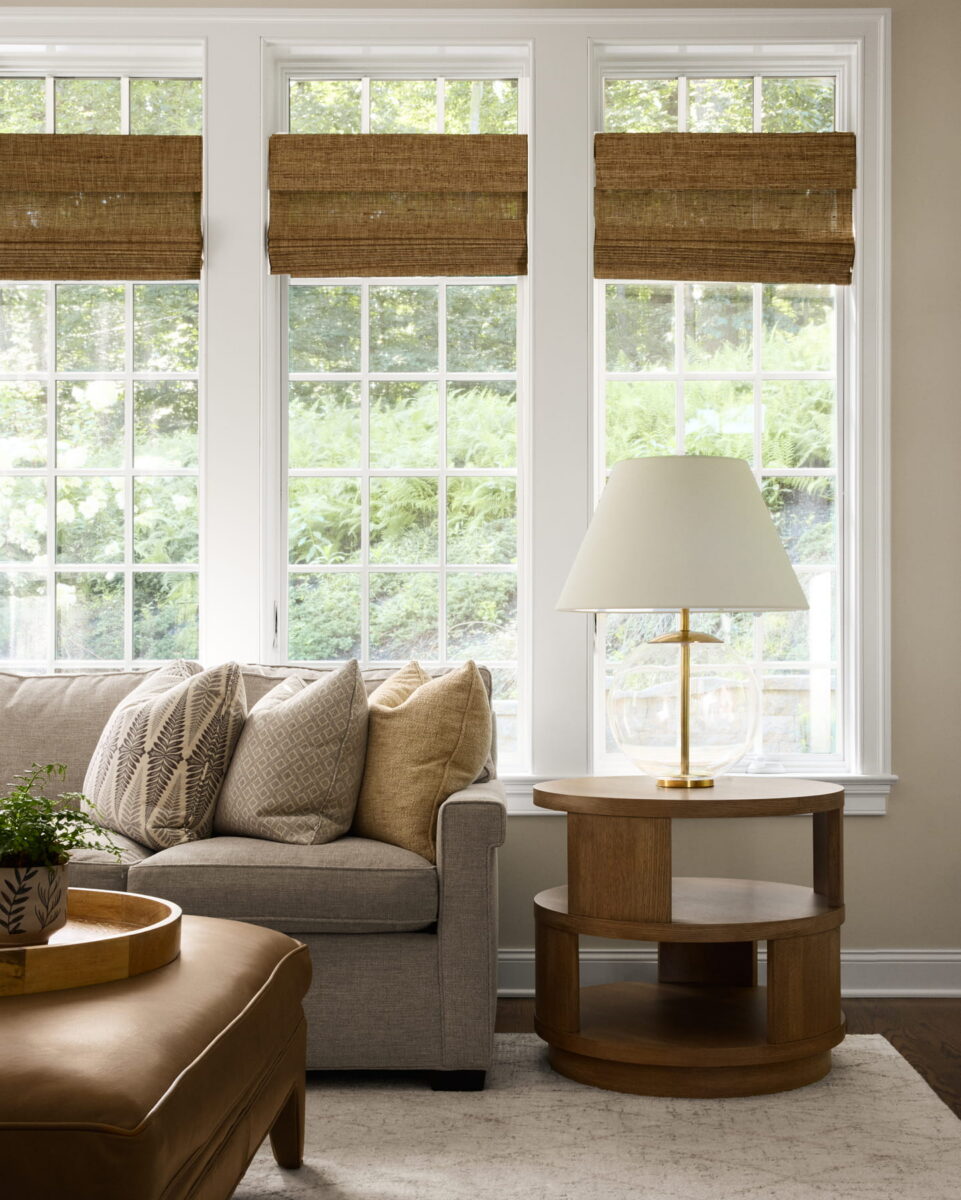A cozy living room with a gray sofa, patterned pillows, round wooden side table, table lamp, potted plant, and large windows with woven shades showing a green outdoor view.