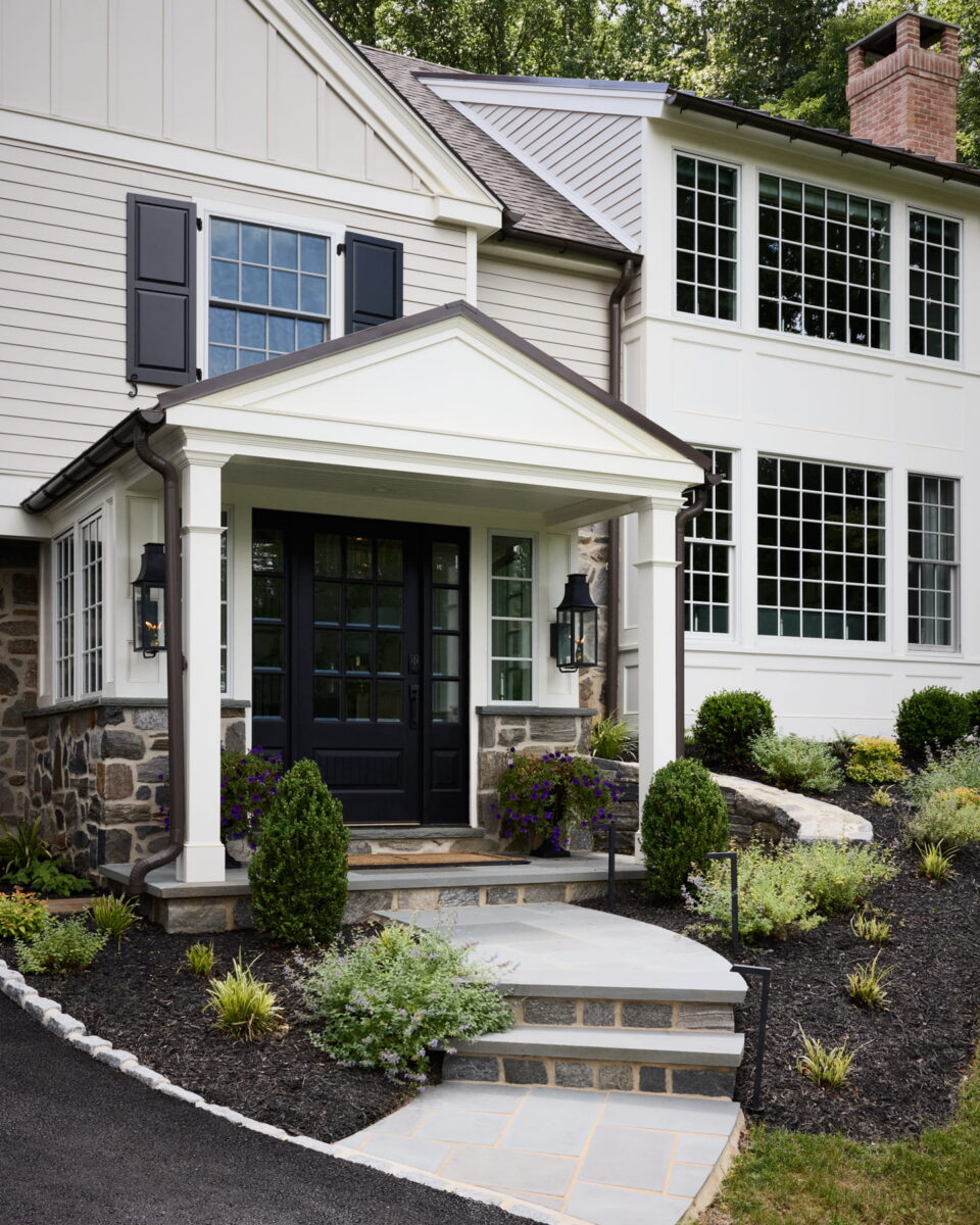 Modern house with white siding, large windows, black front door, and stone accents. Curved stone walkway leads to steps, surrounded by neatly landscaped bushes and mulch.