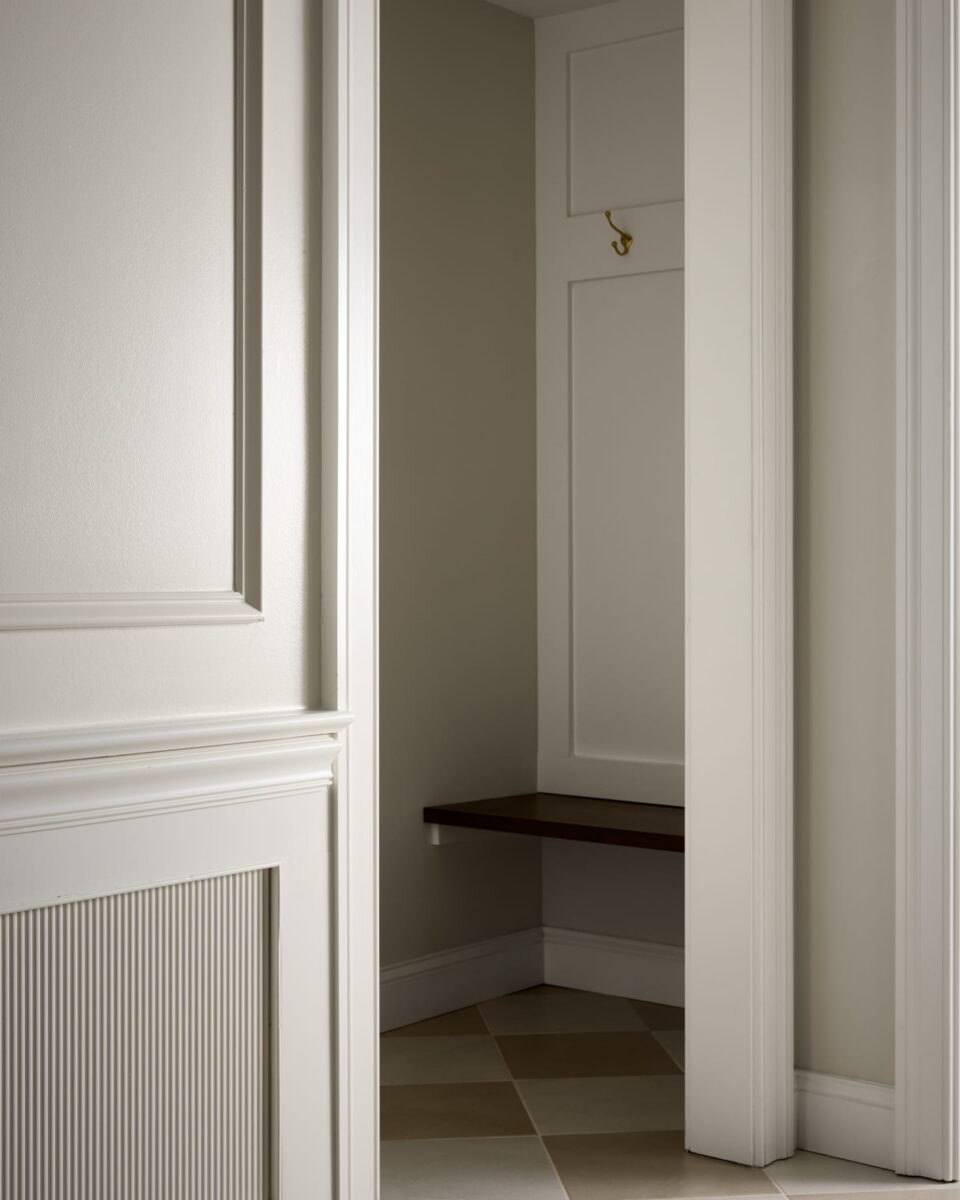 A small, empty nook with beige walls, a single gold wall hook, a dark wooden bench, and checkered tile flooring, partially concealed by a white paneled wall.