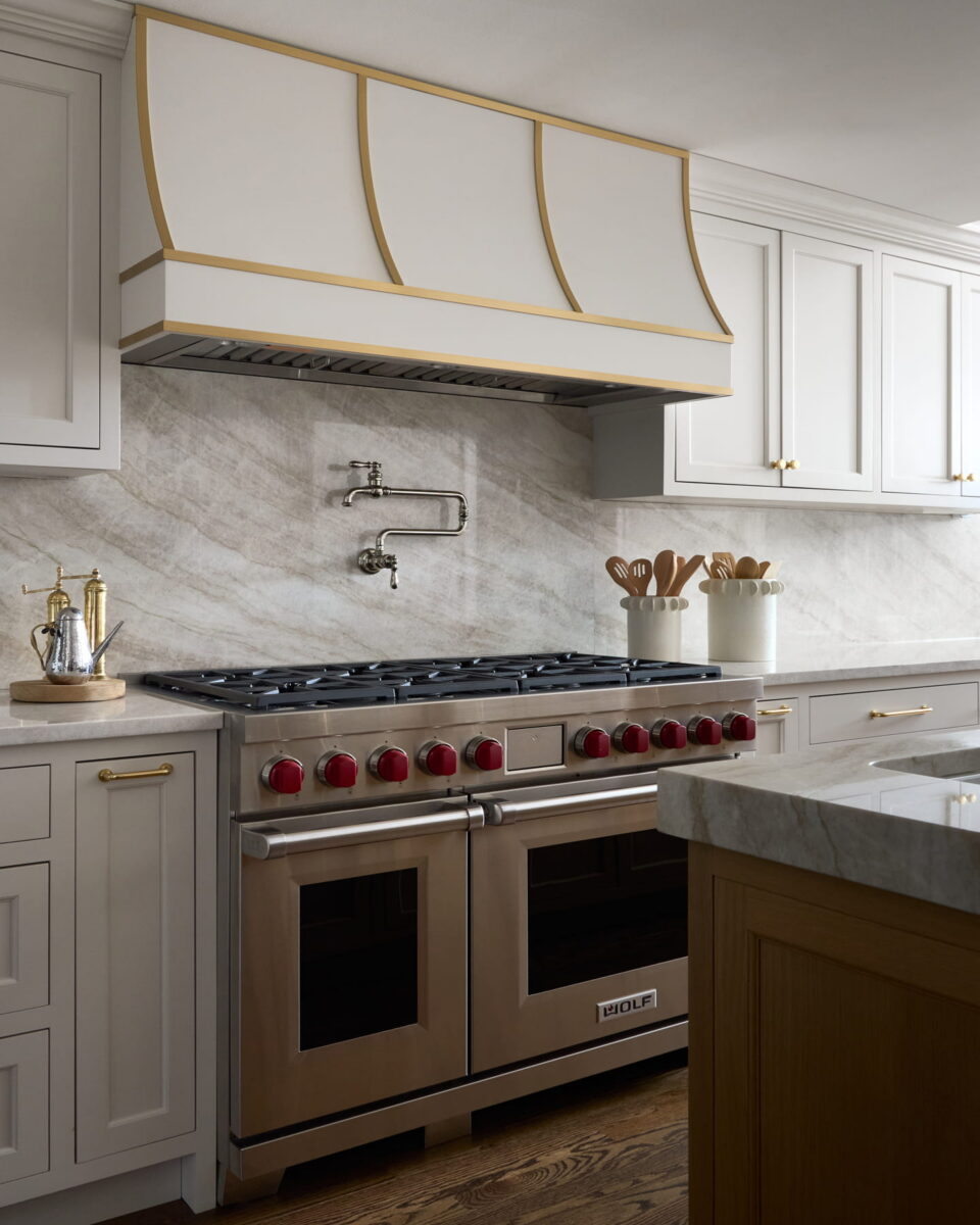Modern kitchen with a stainless steel stove and red knobs, a marble backsplash, white cabinets with gold accents, a pot filler faucet, and utensils in containers on the countertop.
