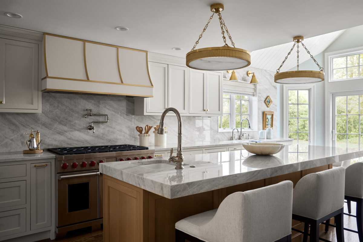 Modern kitchen with marble countertops, white cabinets, gold accents, large island with sink, gas stove, three upholstered bar stools, and large windows letting in natural light.