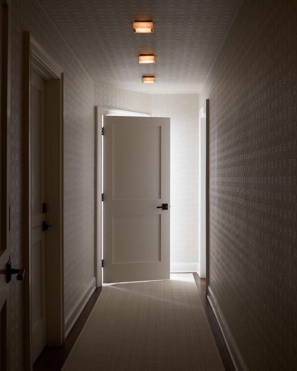 A dimly lit hallway with patterned wallpaper, four ceiling lights, and a light-colored carpet leads to a white door that is open, revealing a brightly lit room beyond.