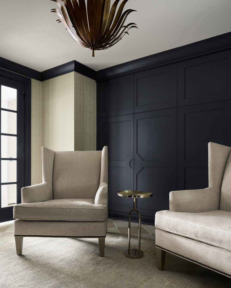 Two beige armchairs and a small round metal side table sit in a modern room with dark paneled walls, light textured wallpaper, a large window, and a decorative brown ceiling light fixture.