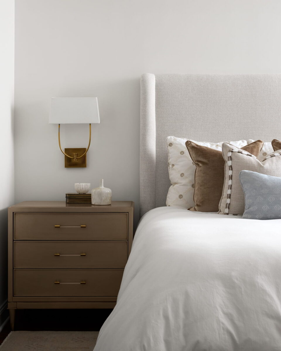 A neatly made bed with neutral pillows beside a beige nightstand holding books, a candle, and a small jar. A modern wall sconce with a white shade is mounted above the nightstand.
