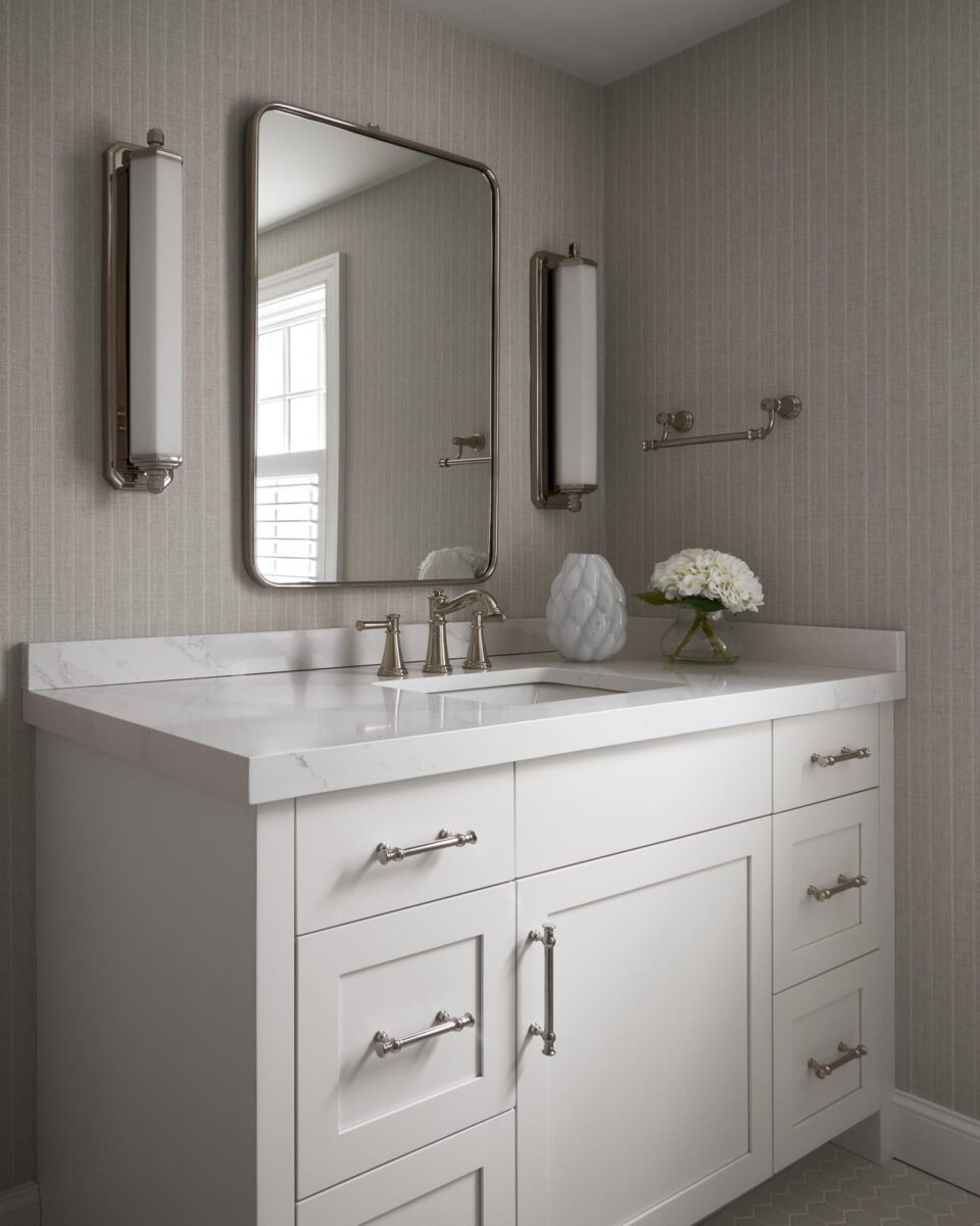 Modern bathroom with a white vanity, marble countertop, rectangular mirror, two vertical lights, a vase with white flowers, and textured wallpaper. A window reflects in the mirror.