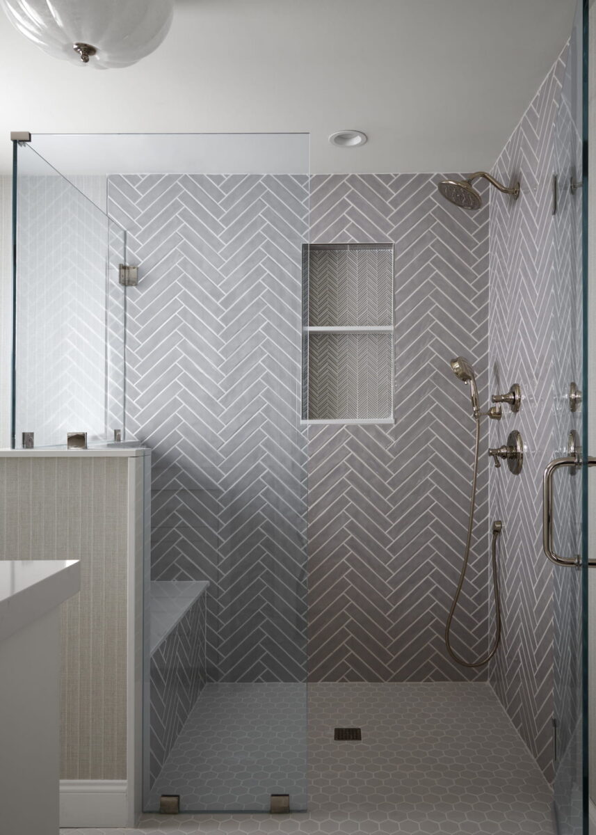 Modern bathroom with a glass-enclosed shower featuring gray herringbone tiles, built-in bench, wall niche, and stainless steel fixtures. The floor is tiled in a small hexagon pattern.