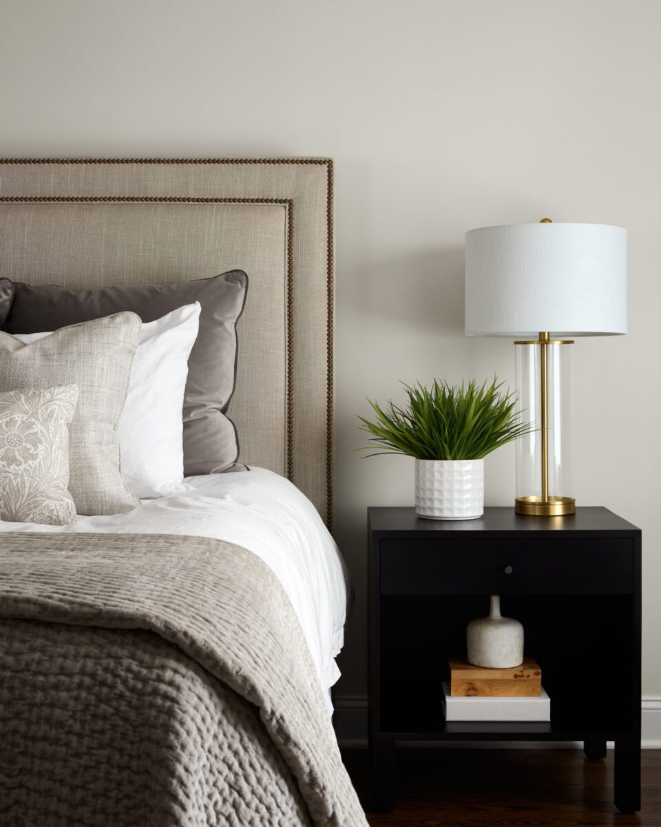 A neatly made bed with neutral bedding and pillows is next to a black nightstand holding a white lamp, a potted plant, and some books against a light-colored wall.