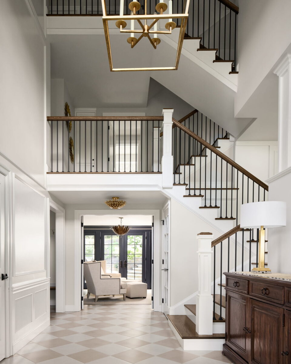 A bright, modern foyer with a multi-level staircase, black railings, a geometric chandelier, checkered tile floor, and a wood console table with a lamp; natural light streams in from the back seating area.
