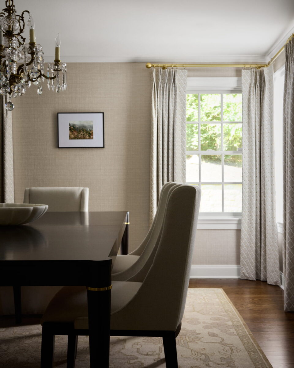 A dining room with a dark table, cream upholstered chairs, a chandelier, a framed picture on the beige wall, and sunlight streaming through a window with patterned curtains.