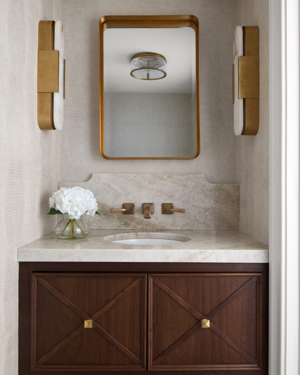 Elegant bathroom vanity with a marble countertop, gold fixtures, rectangular mirror, modern gold wall sconces, brown wood cabinetry, and a glass vase holding white flowers. Ceiling light is reflected in the mirror.