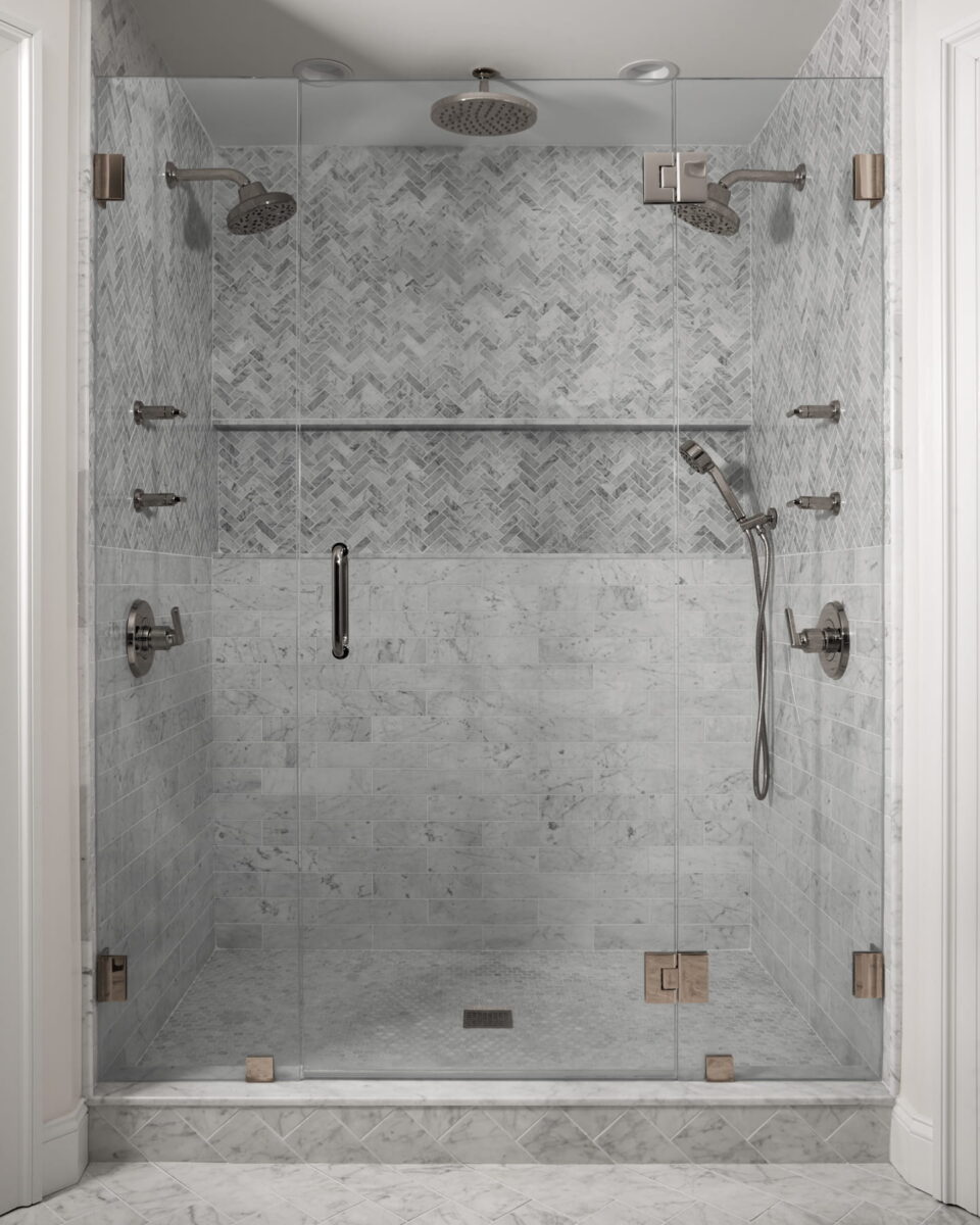 Modern glass shower with gray marble tile, two rainfall showerheads, a handheld shower, and multiple wall-mounted fixtures; herringbone pattern on the upper walls.