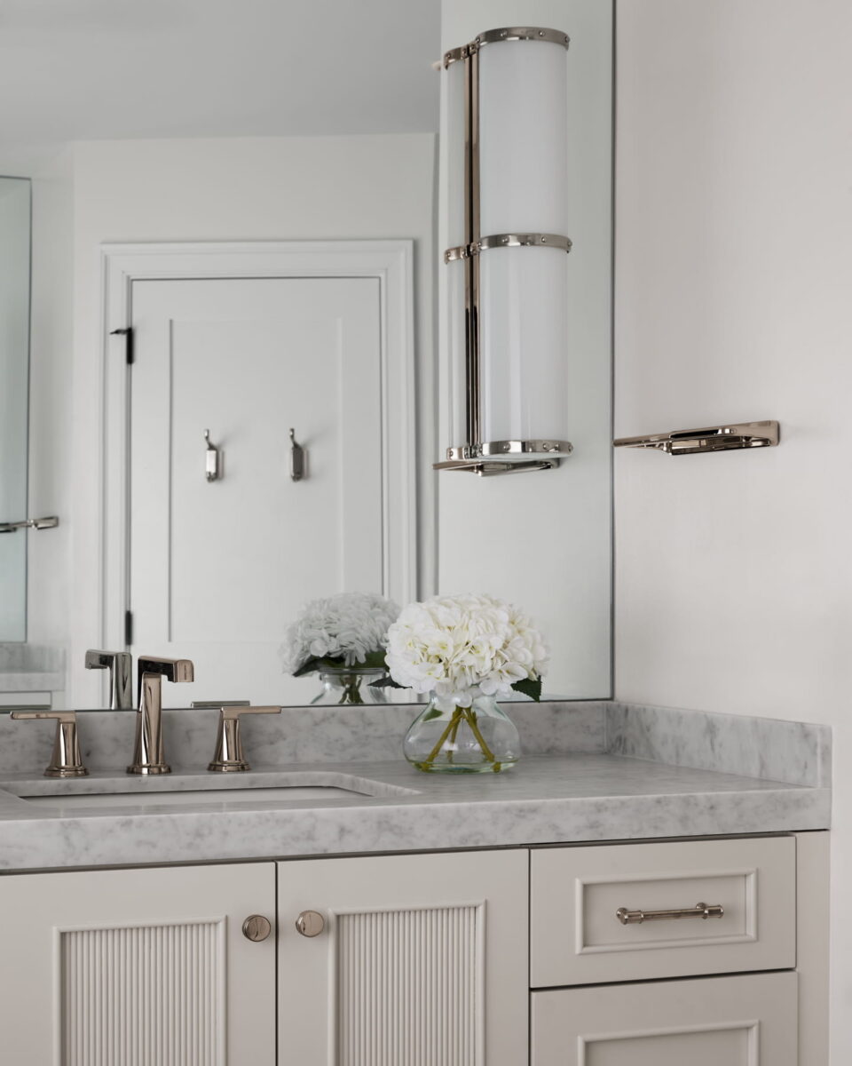 A marble bathroom vanity with a modern faucet, a glass vase of white flowers, a wall mirror, and a tall, cylindrical light fixture. The background shows a white door with two hooks.