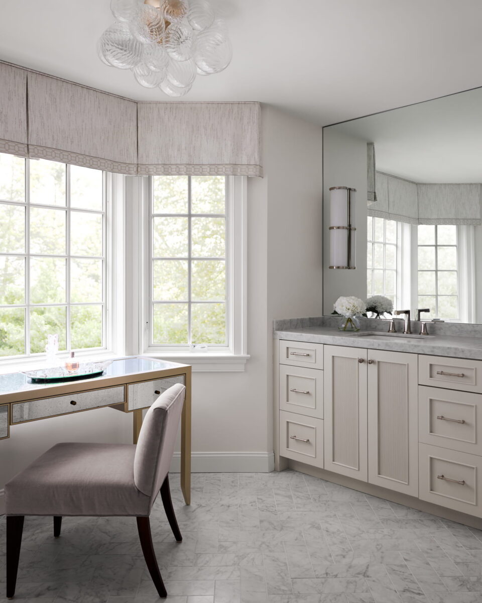 A bright, elegant bathroom features a vanity with a gray chair, marble countertop, large windows with valances, and light cabinetry. A vase of white flowers and toiletries sit on the countertop. Marble floor tiles complete the look.