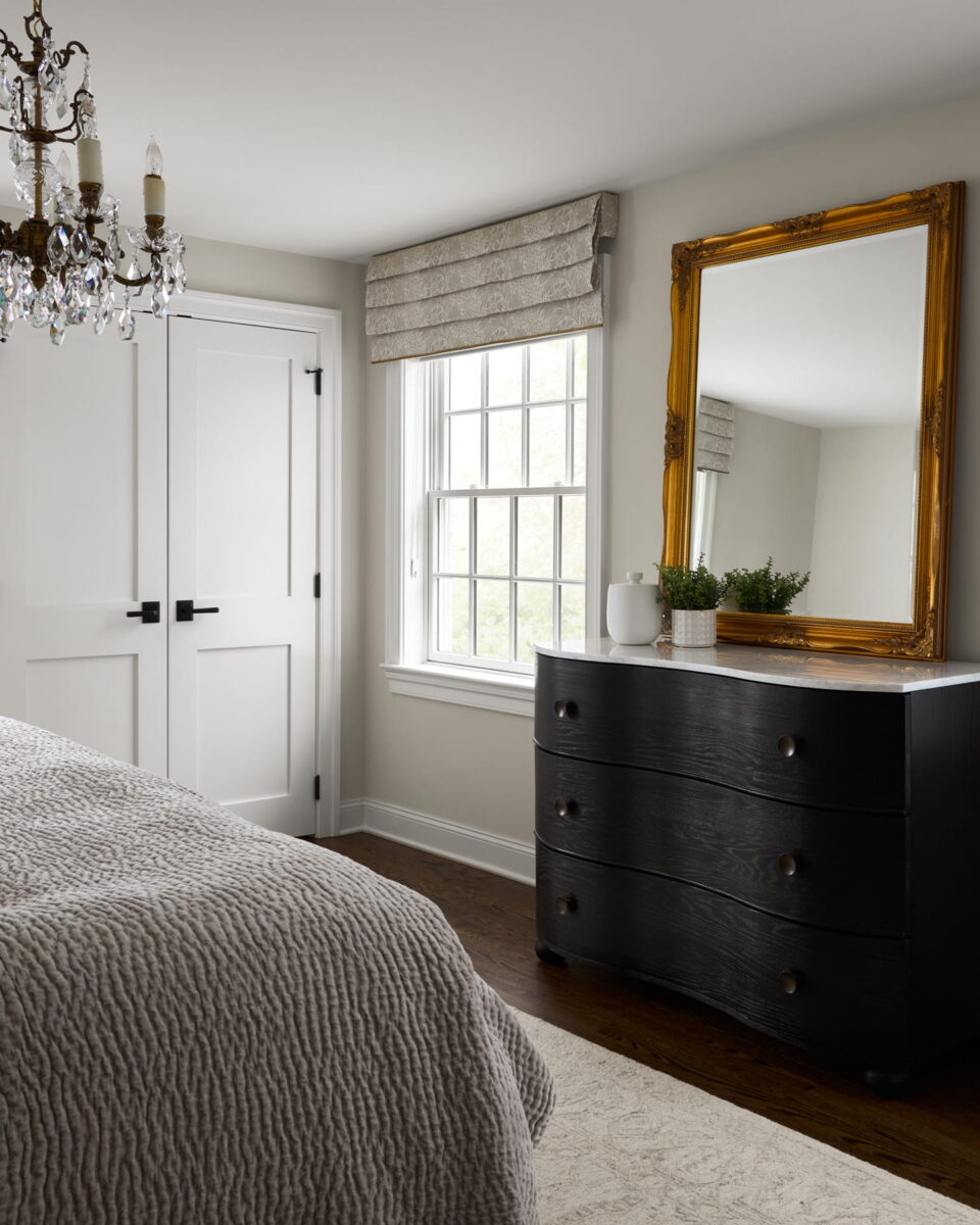 A bedroom with a textured gray bedspread, dark dresser with a gold-framed mirror, a small potted plant, a crystal chandelier, and a window with a Roman shade, letting in natural light.