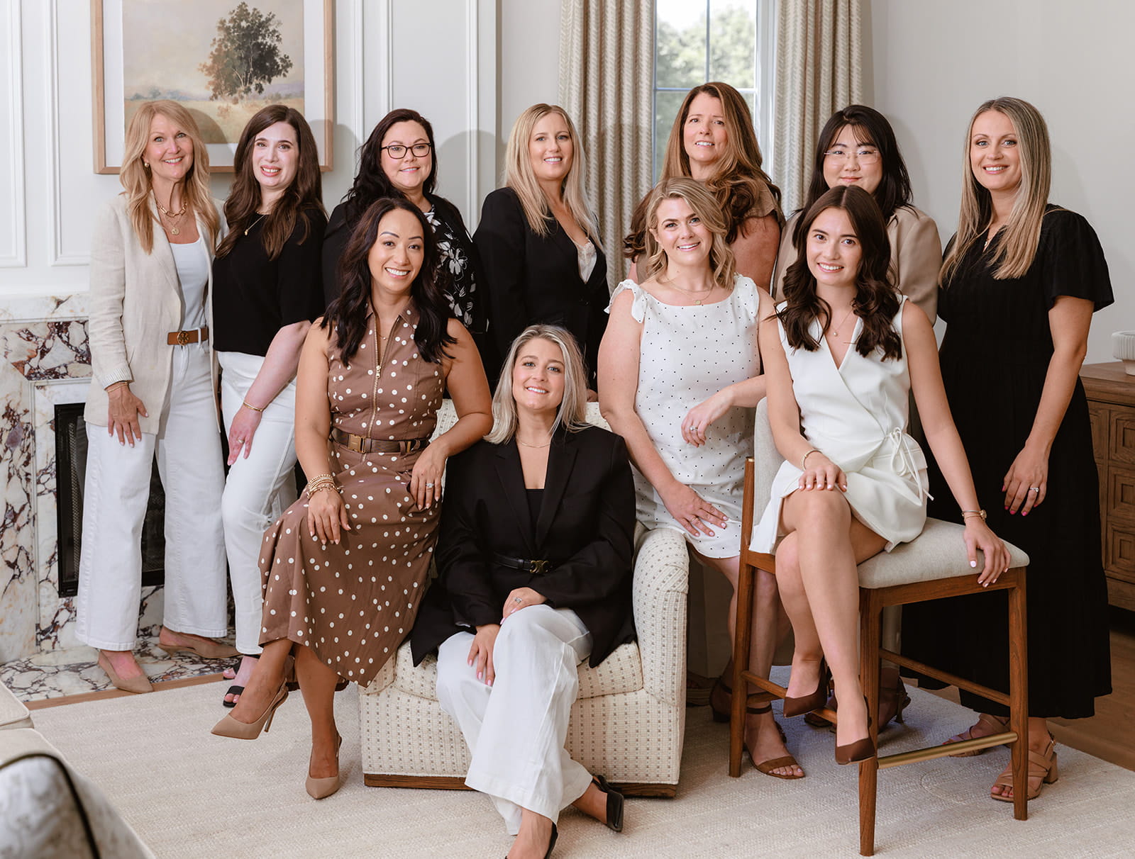 Eleven women pose together in a bright, elegant living room. Some are seated on chairs and a couch, while others stand behind them, all smiling at the camera. The setting appears professional and welcoming, evoking the atmosphere of a vibrant team ready to serve as your design partner for builders.