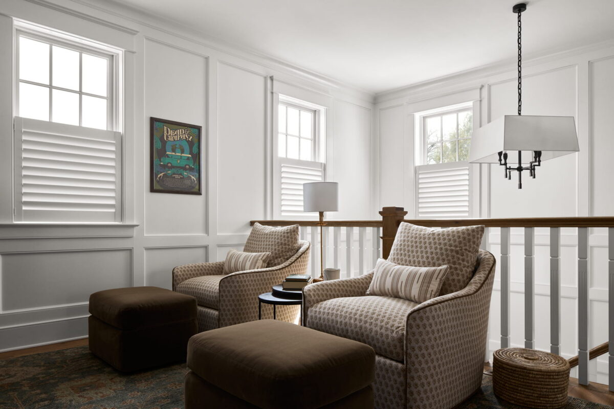 Two patterned armchairs with matching ottomans sit by a wooden railing in a bright room with three windows, white shutters, a floor lamp, side table, woven basket, and a framed vintage painting on the wall.