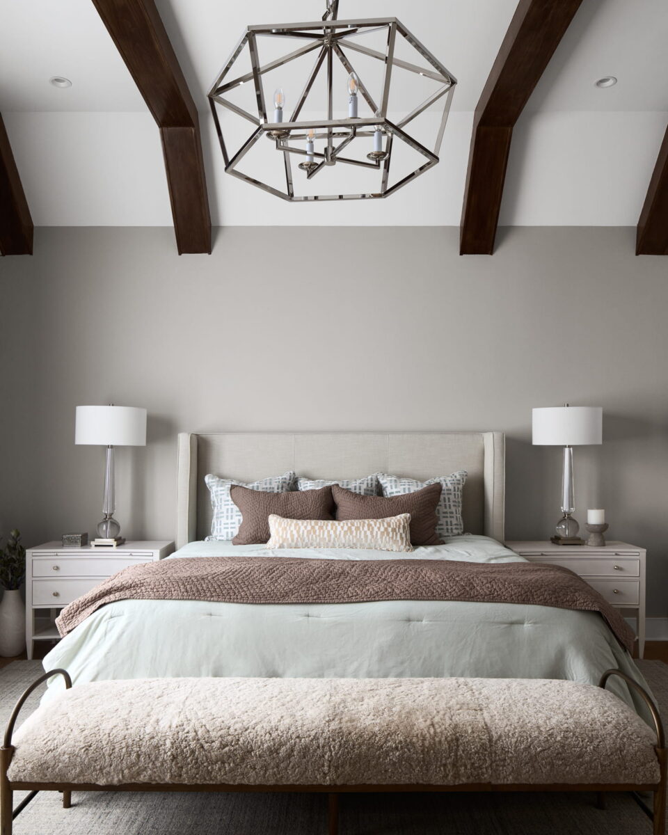 Modern bedroom with a large bed, neutral bedding, two bedside tables with lamps, exposed wooden ceiling beams, a geometric chandelier, and a cushioned bench at the foot of the bed.