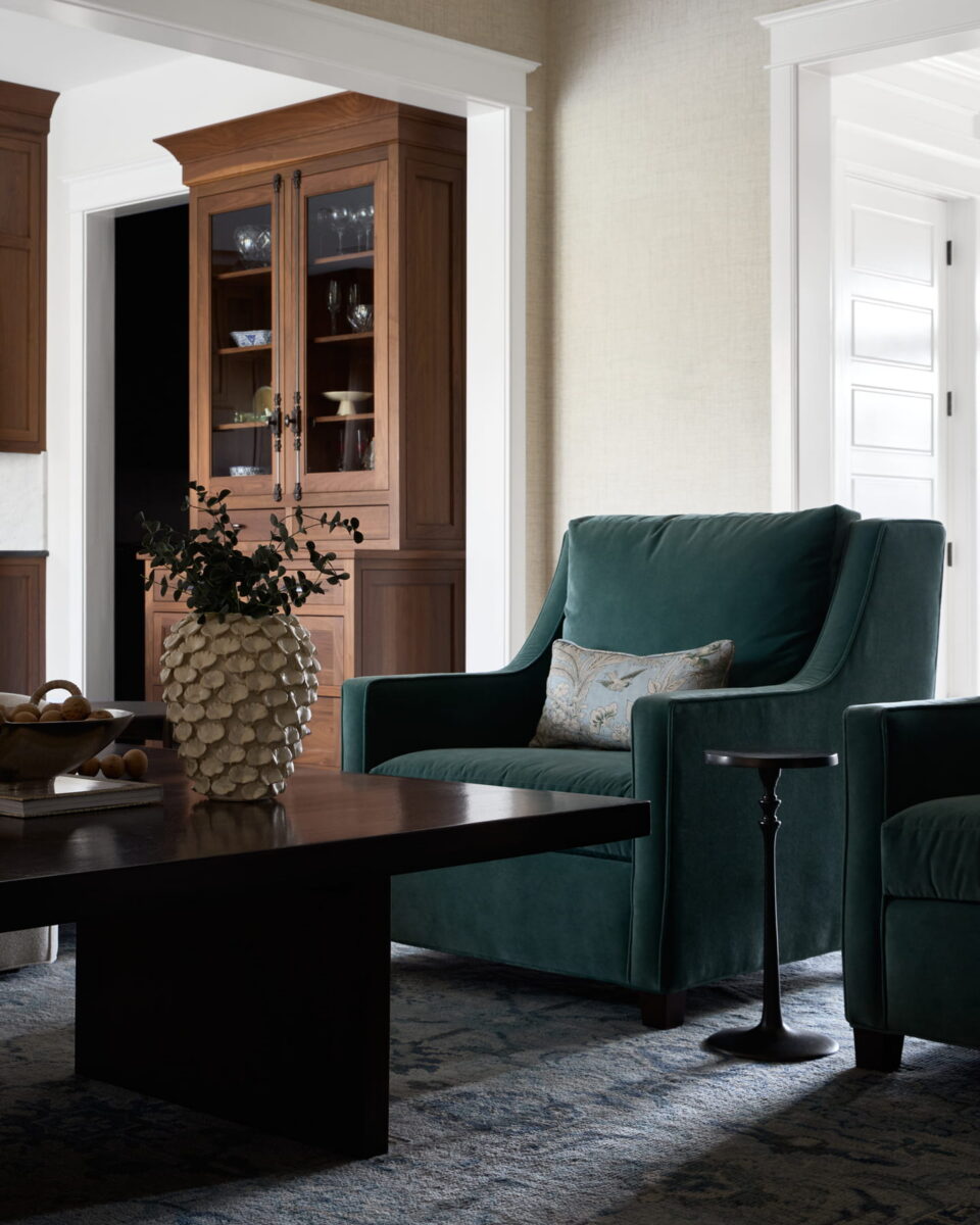 A cozy living room with a green armchair, patterned pillow, dark wood coffee table, textured vase with foliage, and a wooden cabinet displaying dishes in the background. Natural light brightens the neutral walls.