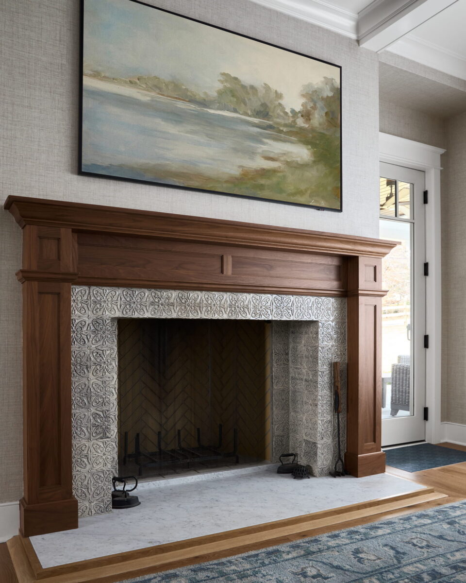 A wooden fireplace with ornate tiles and tools stands below a large landscape painting. Sunlight streams through a glass door on the right. A blue rug covers the wooden floor.